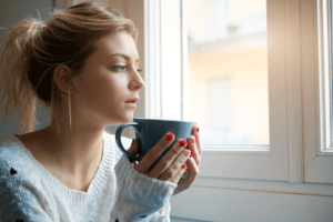 young woman sipping coffee while looking out window and wondering how to manage emotions in sobriety