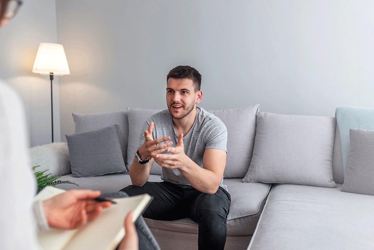a man sits on a couch and is talking to his therapist while she takes notes and explains to him alcohol detox benefits