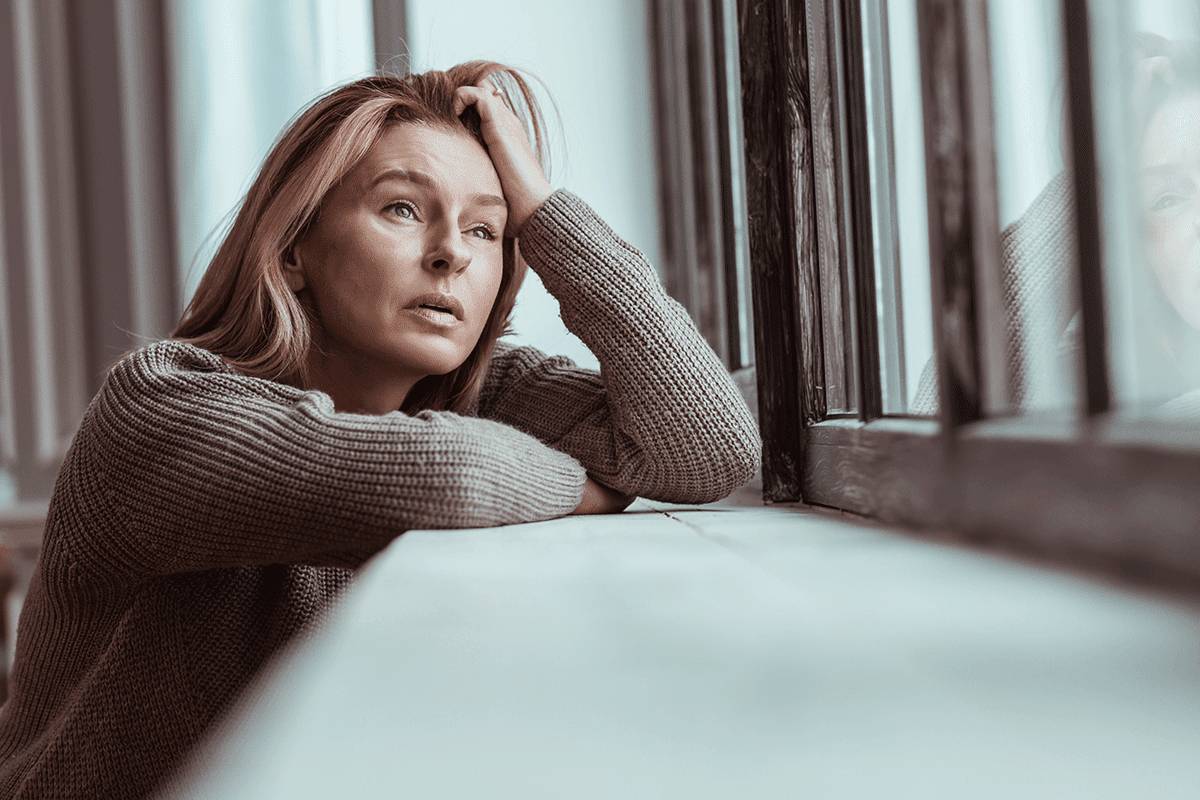 woman leans on a window ledge with her head resting on her arm while thinking about the signs of alcoholism