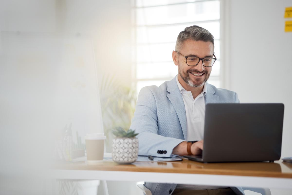 man with glasses sits a desk smiling and typing on laptop while researching the benefits of living substance free