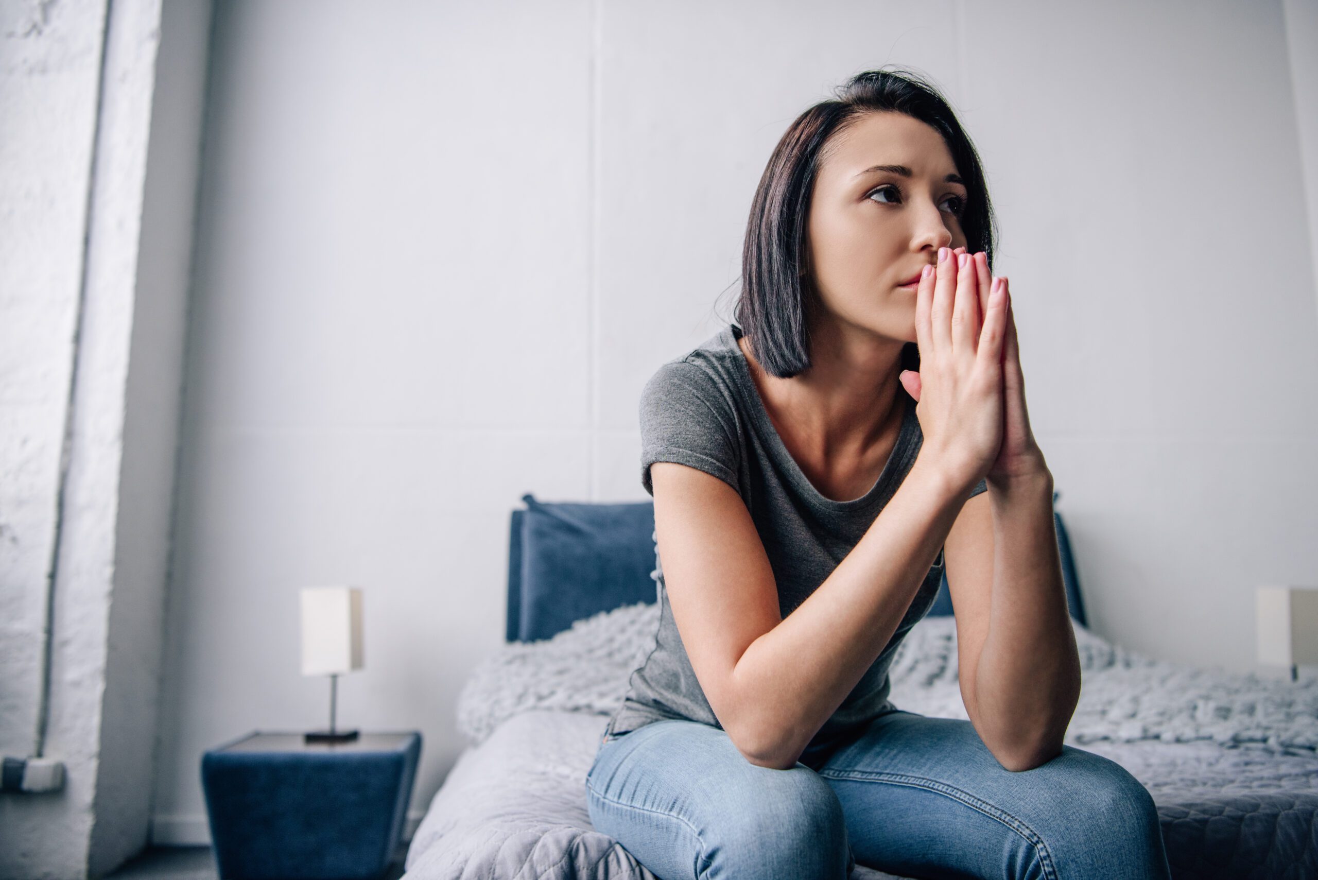 woman sits on the edge of her bed with her palms together and thinks about what are the signs that someone is using drugs