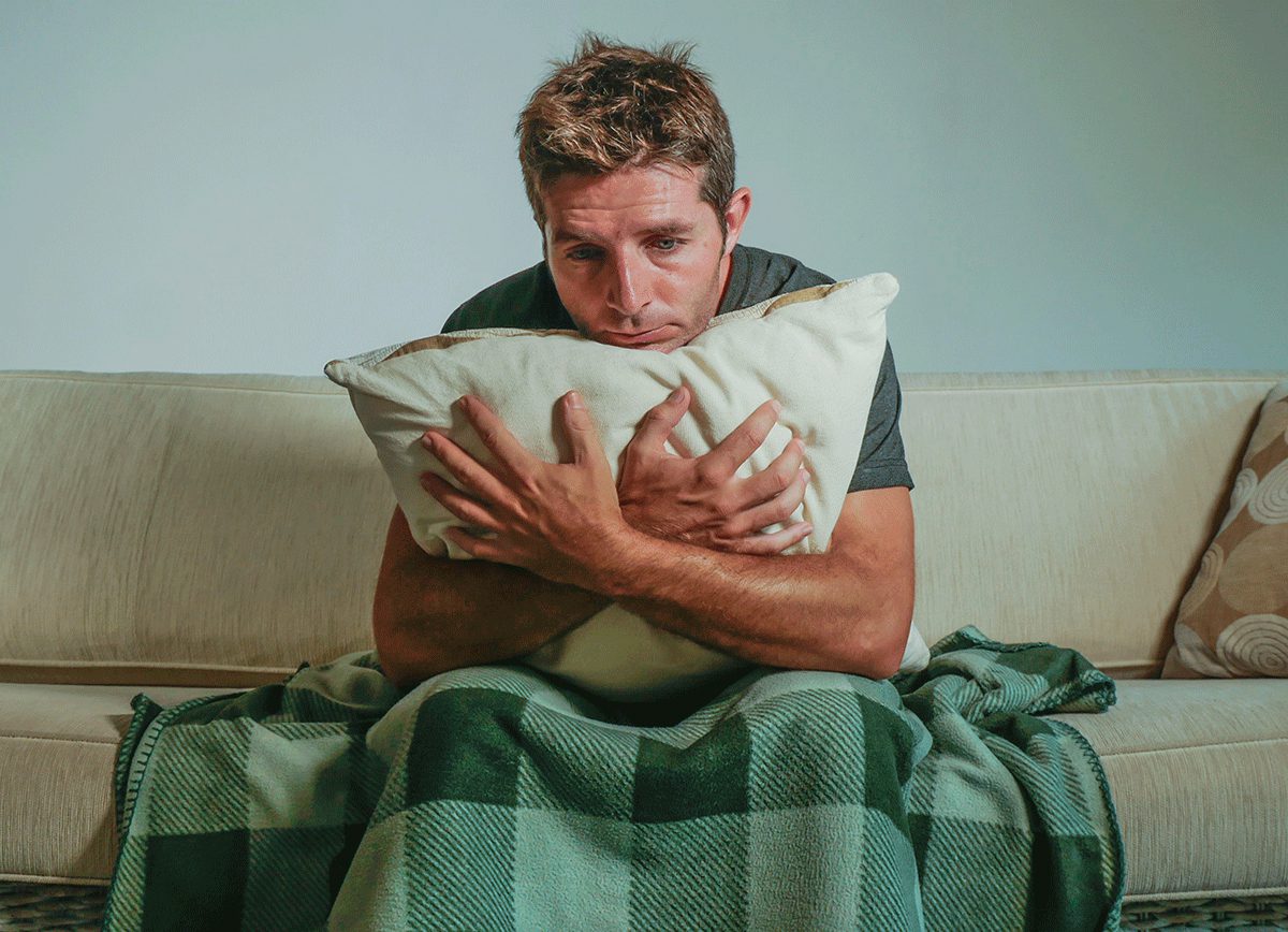 man hugs a pillow while sitting on his couch with a blanket and experiencing cocaine withdrawal